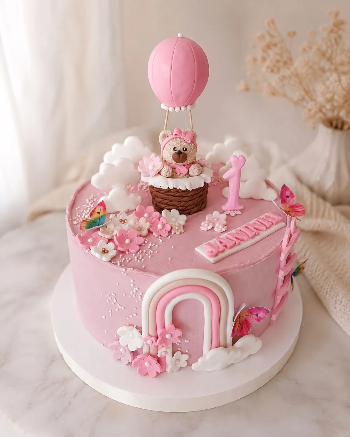 Różowy tort na roczek z pluszowym misiem w wiklinowym koszyku balonu, pastelową tęczą i różową jedynką