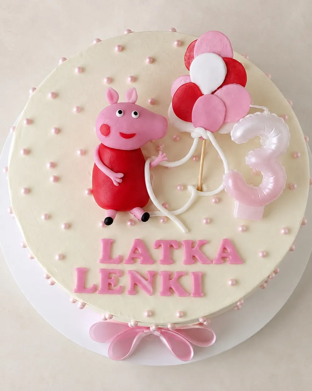 Tort z góry: figurka świnki obok pęku balonów, napis Latka Lenki i różowa wstążka u podstawy